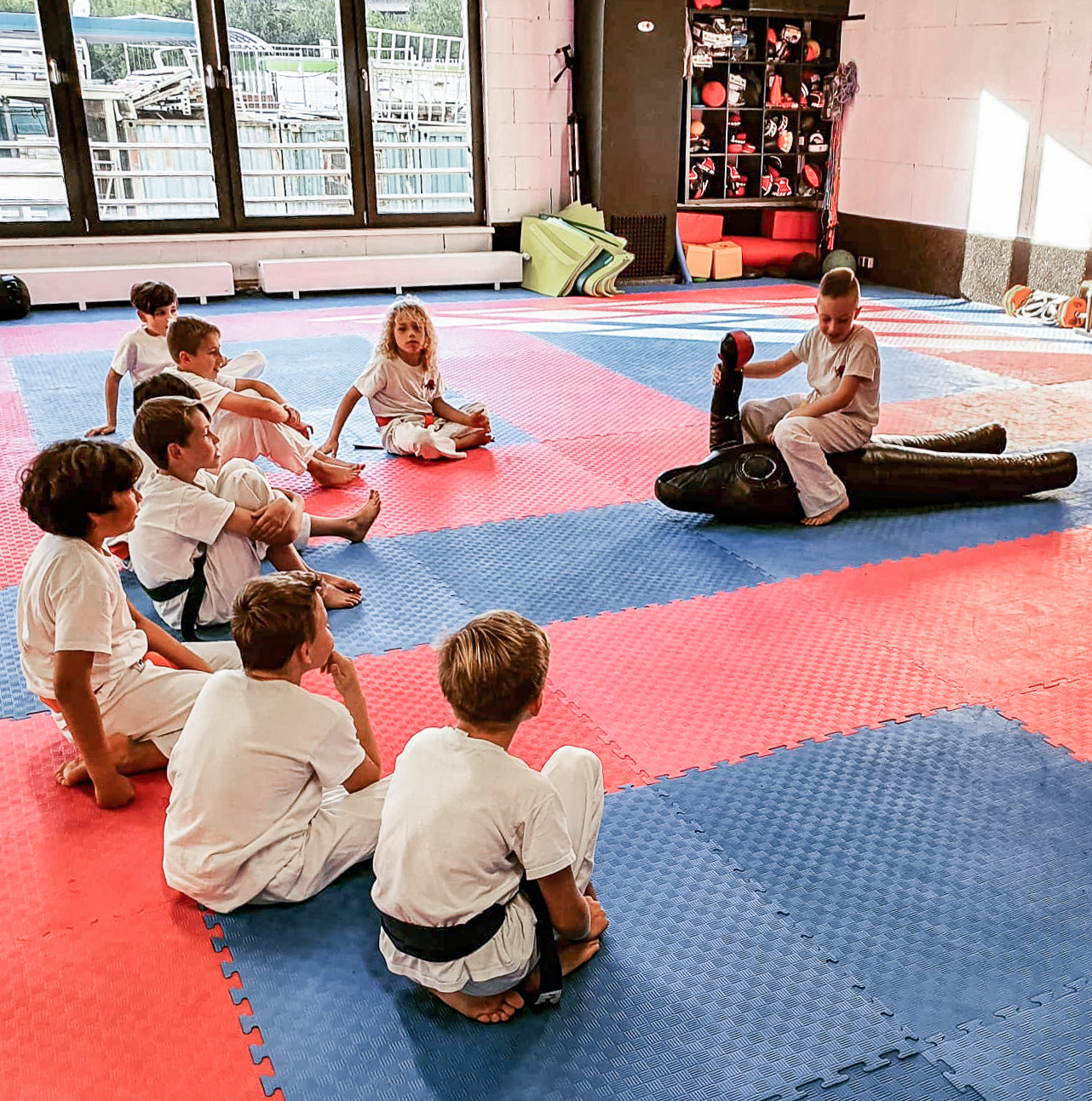 Kinder sitzen in einem Halbkreis um einen Jungen, der gerade mit einer Trainingspuppe trainiert